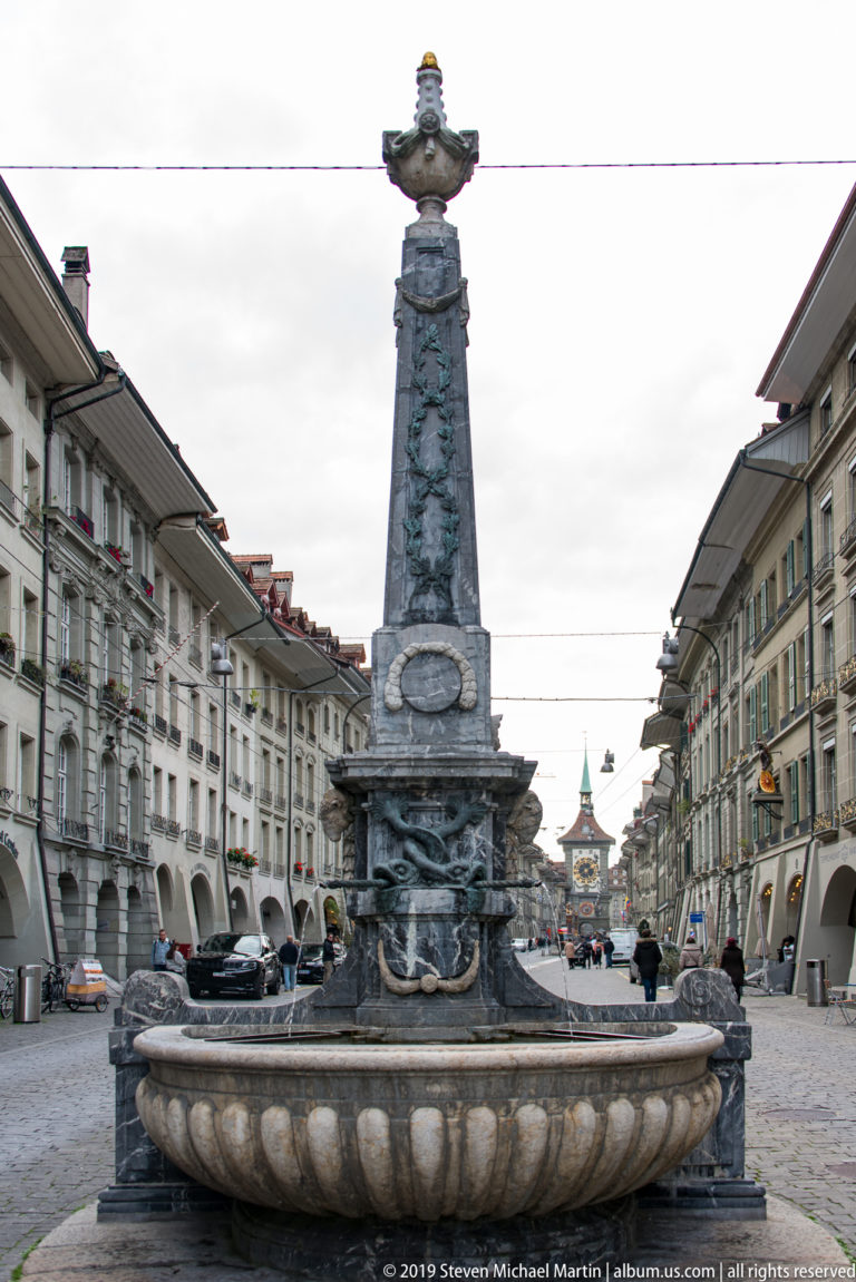 Large stone street fountain with tall sculpted column centered between arcaded historic buildings in Bern.