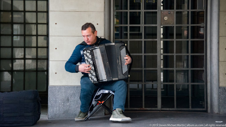 Seated street musician playing an accordion beneath a stone arcade beside a dark doorway.