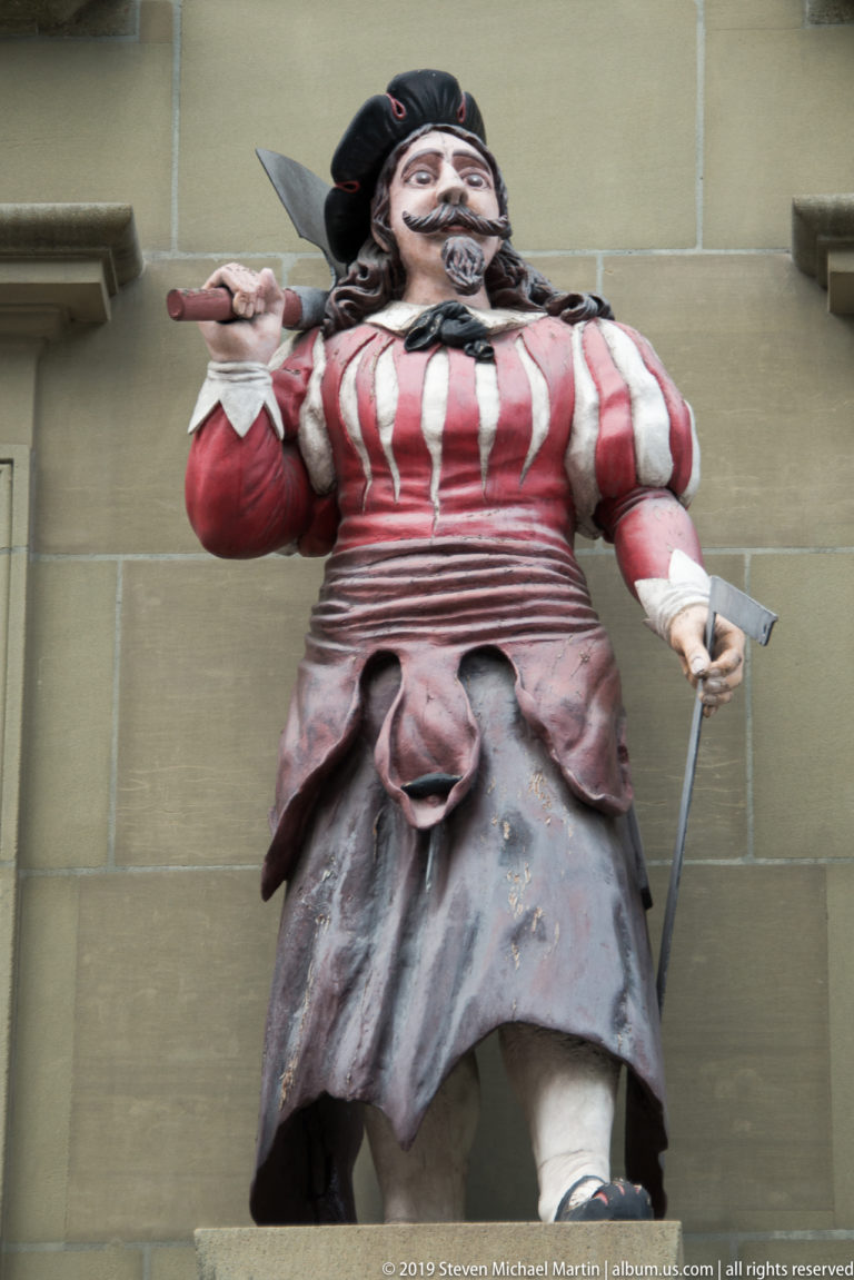 Painted bearded fountain figure in red-and-white striped clothing holding long implements against a stone facade.