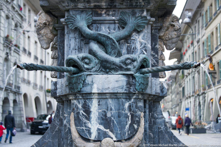 Close detail of a carved fountain face with projecting water pipe and dark sculptural ornament, street blurred behind.
