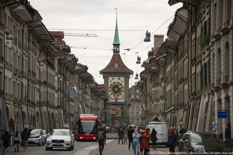 Zytglogge clock tower centered at the end of a busy Bern street with red bus, cars, pedestrians, and arcaded facades.