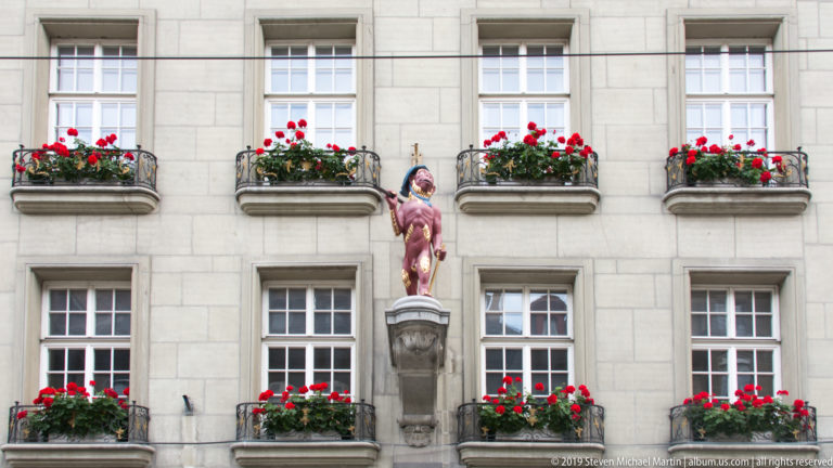 Painted standing fountain figure centered on a column before a pale facade with rows of red flower boxes.