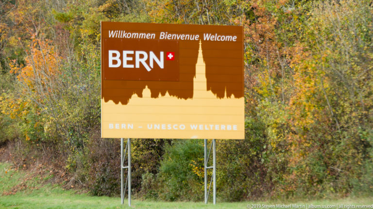 Brown roadside sign reading BERN and “Bern – UNESCO Welterbe” against autumn foliage.