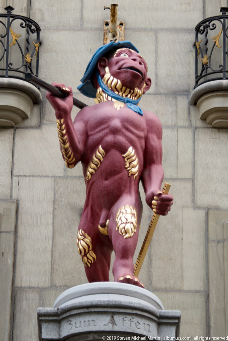 Bright red grotesque or jester-like fountain figure with gold details, extended hand, and sword-like implement.