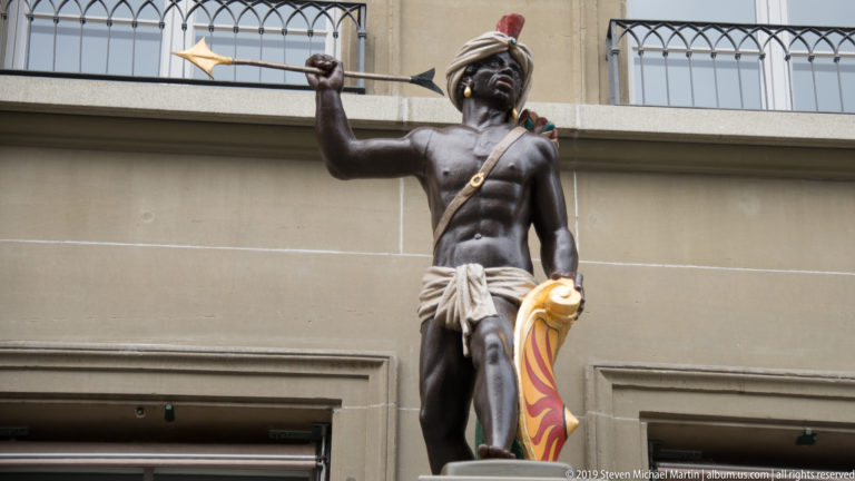 Dark-skinned male fountain figure holding a spear and colorful shield, wearing light drapery and a crested headpiece.