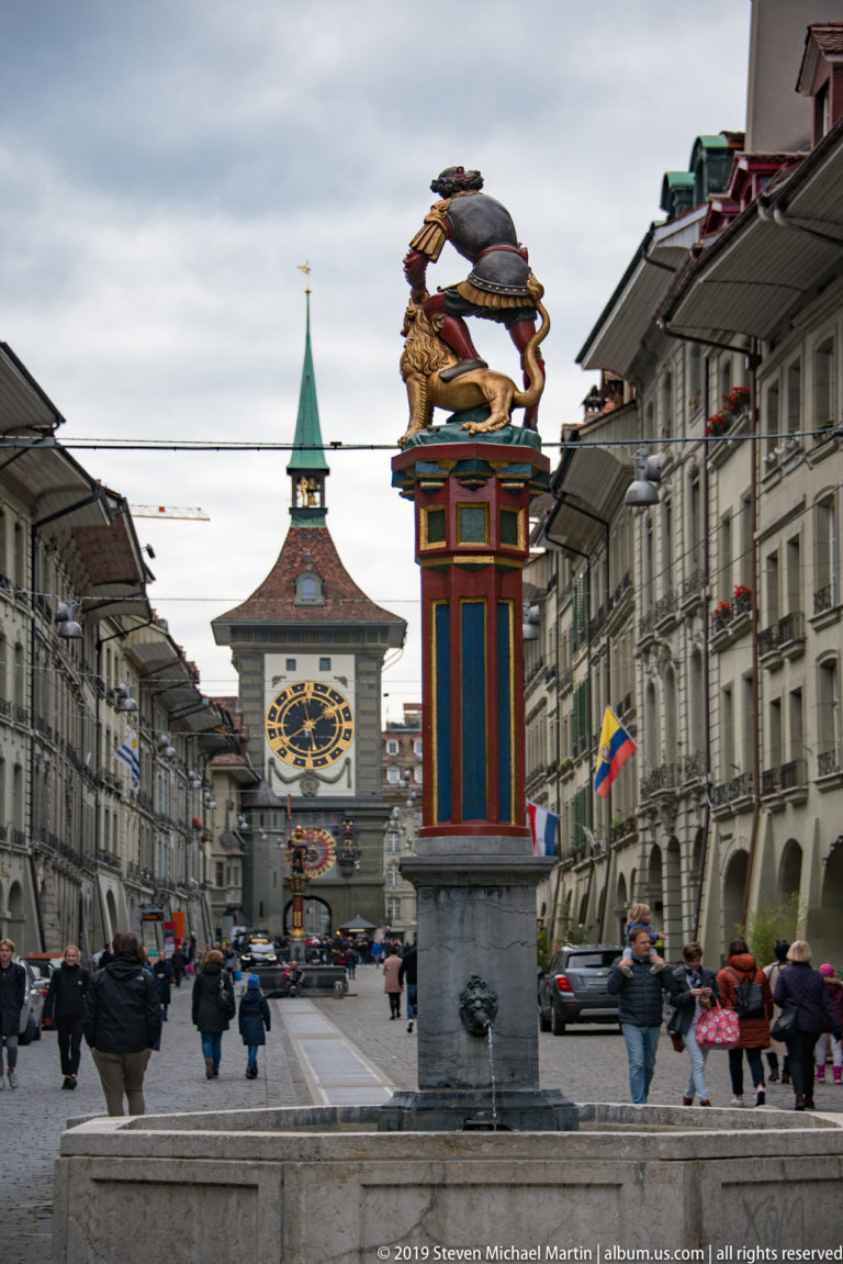Painted fountain column in the foreground with pedestrians and the Zytglogge clock tower aligned in the distance.