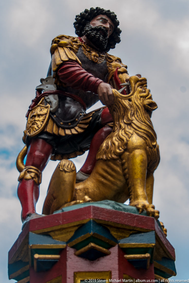 Bearded man in red-and-gold clothing wrestling a golden lion atop a Bern fountain.