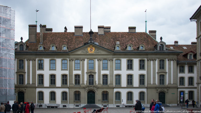 Symmetrical pale Baroque façade with tiled roof, dormers, chimneys, and a central pediment on a Bern square.