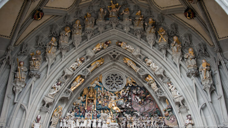 Close view of Bern Minster’s Last Judgment portal with gilded figures arranged around pointed Gothic archivolts.