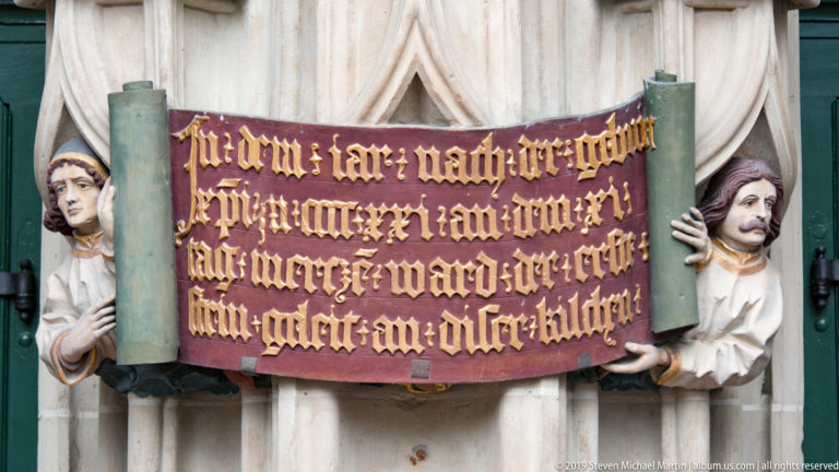 Two sculpted figures holding a red scroll with gilded Gothic-script inscription on Bern Minster’s west portal.