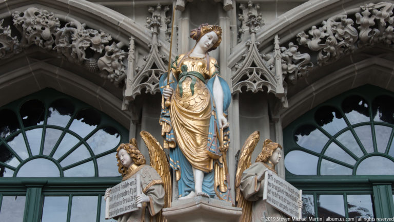 Crowned polychromed female figure above two angels in the sculptural program of Bern Minster’s west portal.