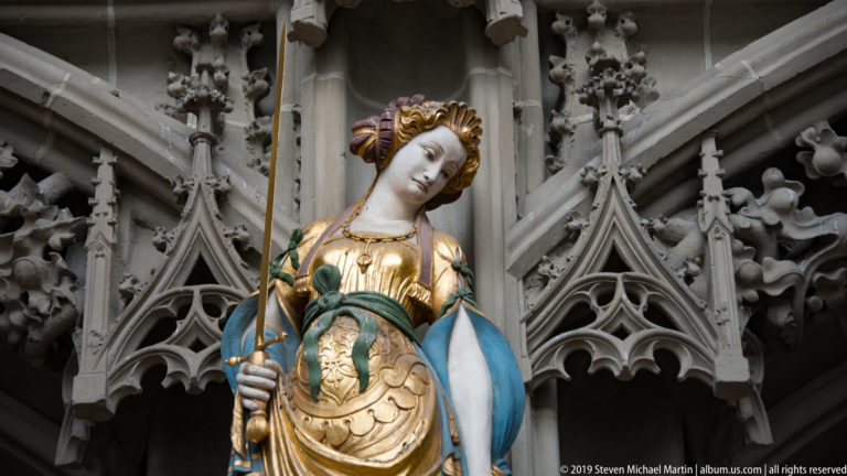 Close view of a crowned polychromed female sculpture with gilded garments on Bern Minster’s west portal.