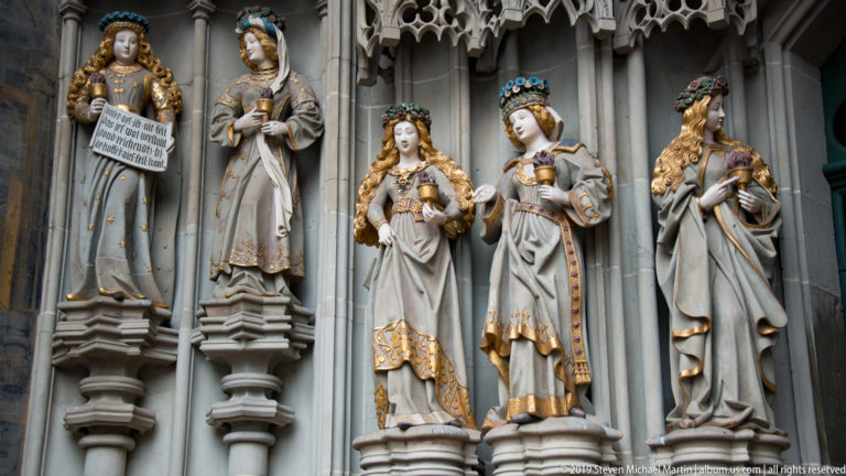 Three richly dressed female sculptures with crowns and gilded details in niches at Bern Minster.