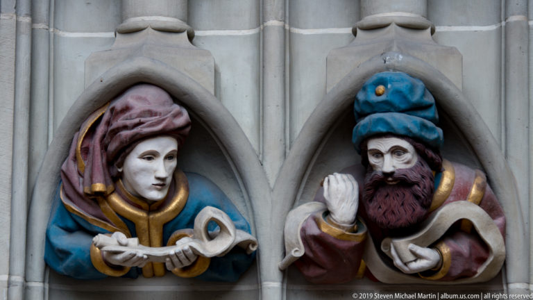 Two polychromed bust figures with elaborate head coverings and scrolls set into pointed stone niches.