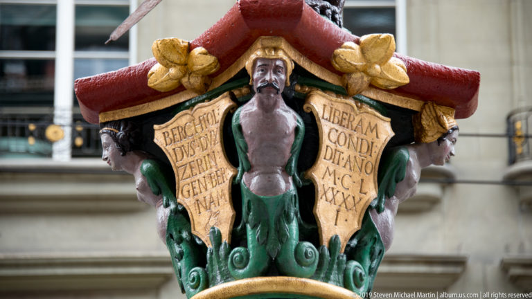 Painted and gilded fountain capital with a small male figure and two inscribed panels on the Zähringerbrunnen.