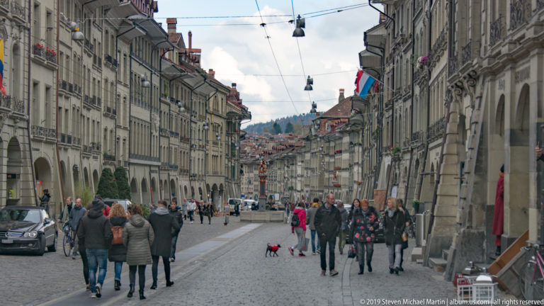 Pedestrians walking along a stone-paved Bern Old City street lined with arcaded historic façades.