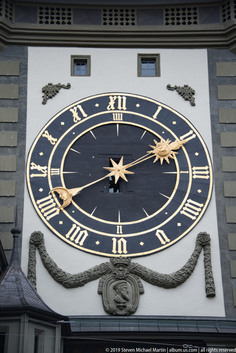 Large black-and-gold Zytglogge clock face with Roman numerals and sun-and-moon motifs.