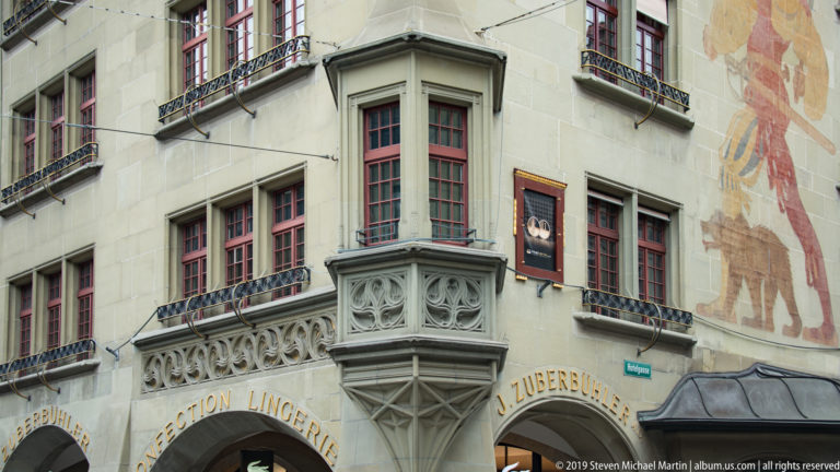 Historic Bern building with carved corner oriel, red-framed windows, mural painting, and gold lettering.