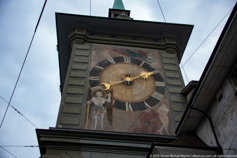 Large historic clock face with gilded celestial symbols and painted figures on a Bern tower façade.