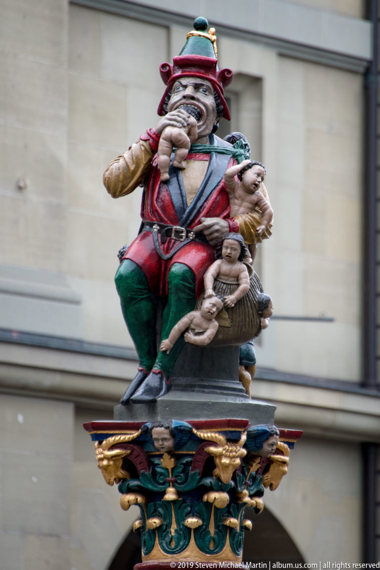 Colorful seated bagpiper fountain figure with a small companion on an ornate gilded capital.