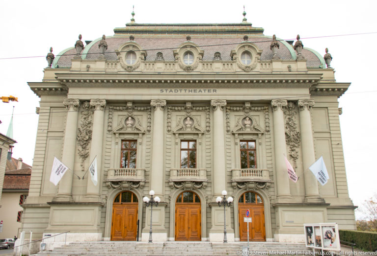 Frontal view of Stadttheater Bern with three wooden entrances and an ornate symmetrical stone façade.