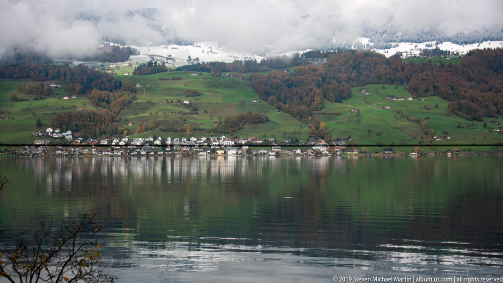 Calm lake reflecting a shoreline village beneath green pastures, autumn woods, cloud, and patches of snow.