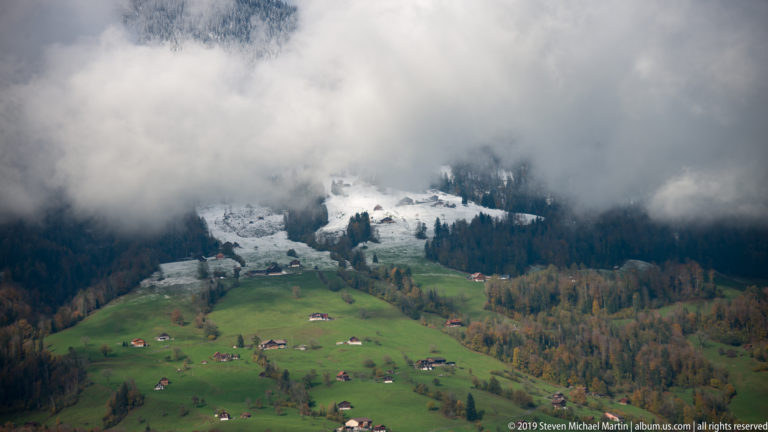 Green Alpine pasture with scattered farm buildings below snowy upper slopes and low cloud.