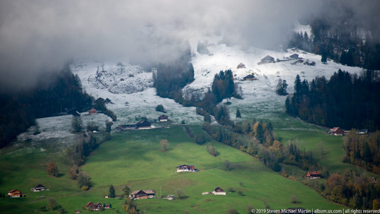 Snow-covered upper hillside descending toward green meadows with scattered Alpine farmhouses and dark conifers.
