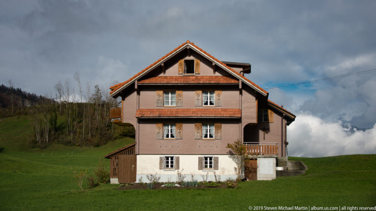 Traditional multi-story Alpine farmhouse with timber upper walls and balconies standing on a green hillside.