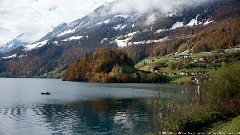 Small boat on an Alpine lake with autumn forest, green shore, and snow-streaked mountains beyond.
