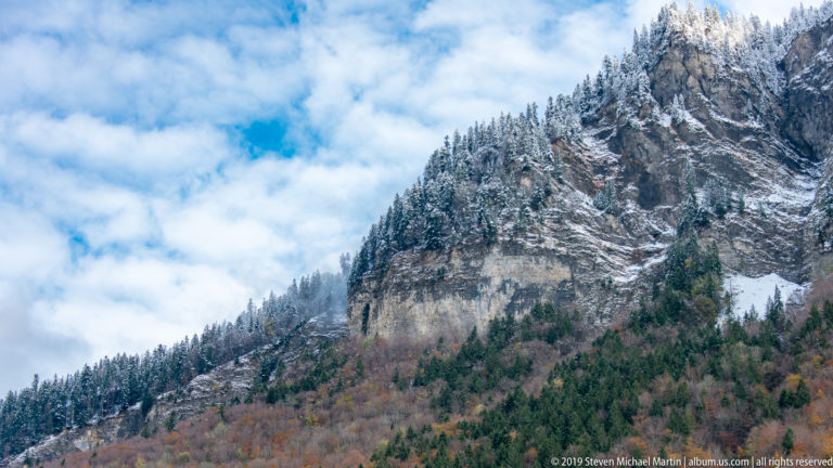 Steep layered rock cliff topped with snow-dusted conifers above autumn woodland under broken cloud.