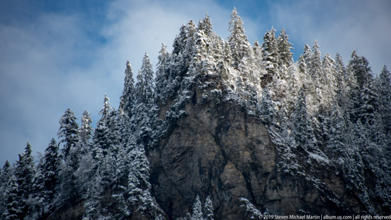 Rugged rock pinnacle crowned and surrounded by snow-covered conifer trees beneath a blue-gray sky.