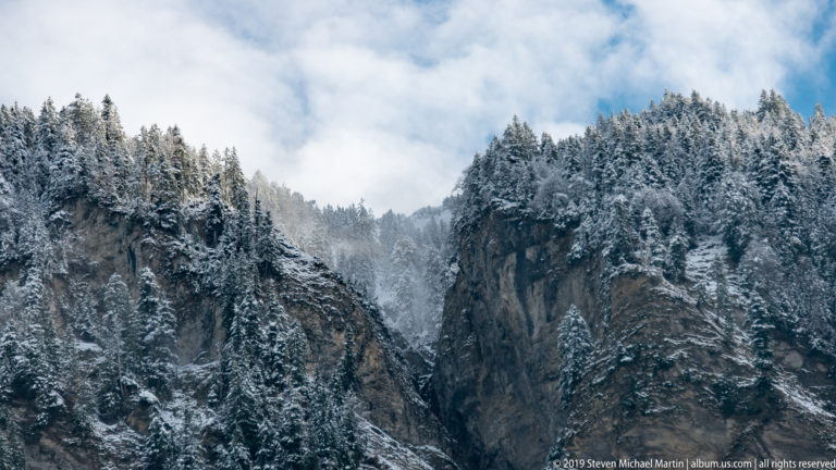 Narrow rocky cleft between steep snow-covered conifer slopes, with mist rising through the gap.