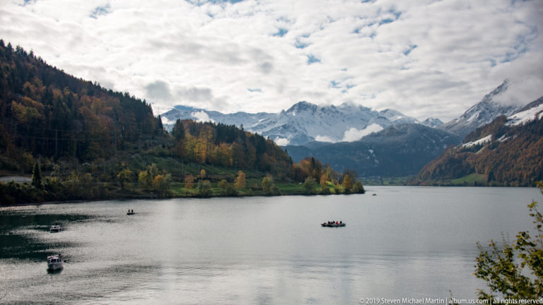 Several small boats on a broad Alpine lake surrounded by autumn slopes and distant snowy mountains.