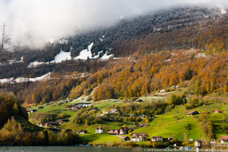 Sunlit green hillside with scattered chalets below autumn forest, snow patches, and low cloud.