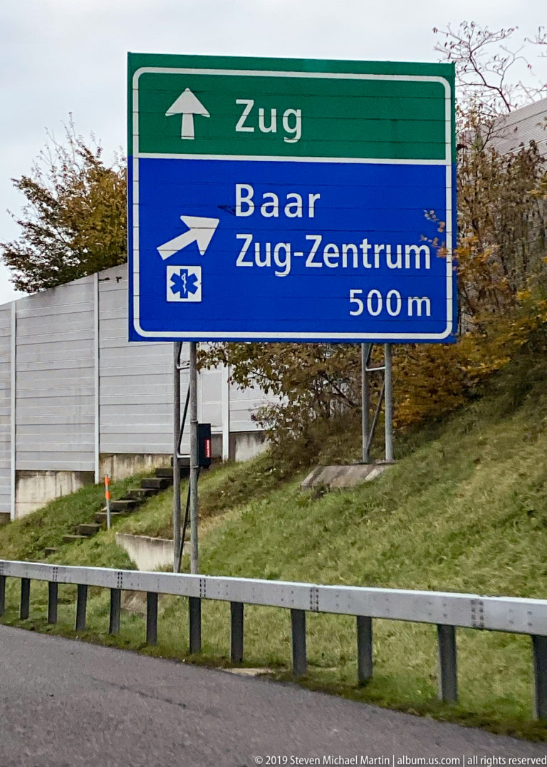 Swiss road signs directing traffic toward Zug, Baar, and Zug-Zentrum beside a highway guardrail.
