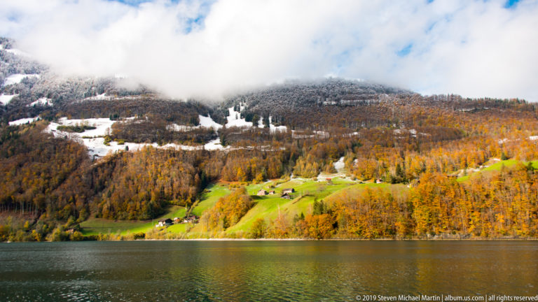 Dark lake below a hillside of golden autumn forest, green meadows, snow patches, and low cloud.
