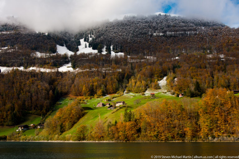 Green farm clearing and small buildings above a lake, surrounded by autumn forest with snow higher on the slope.