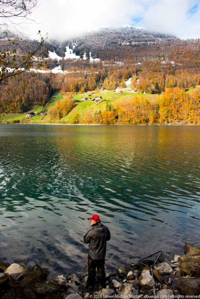 Angler in a red cap fishing from a rocky Alpine lakeshore, with autumn hills and snow across the water.
