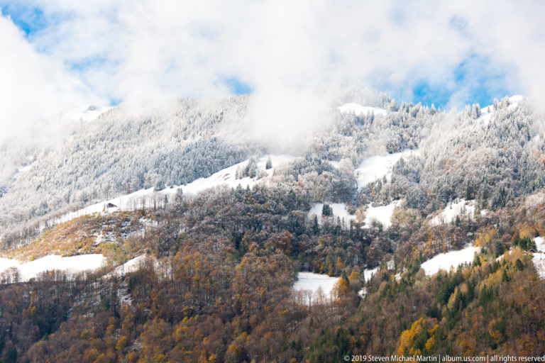 Forested mountainside mottled with fresh snow, autumn foliage, open clearings, and low cloud.