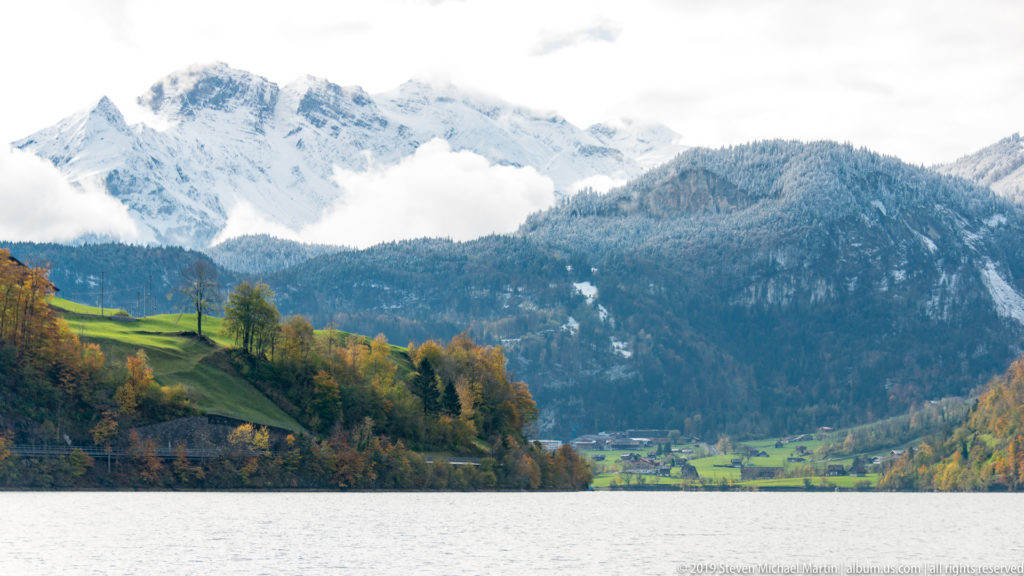 Alpine lake leading toward a green valley beneath forested slopes, snowy mountains, and drifting cloud.