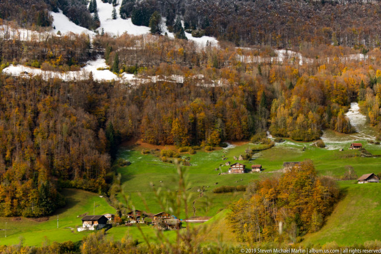 Scattered chalets in bright green Alpine meadows surrounded by autumn forest and bands of snow.