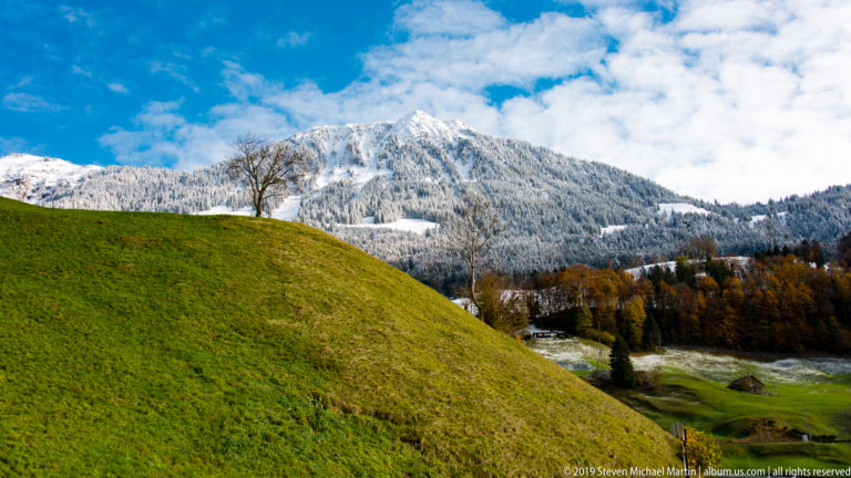 Bright green pasture mound in front of a snowy forested mountain ridge beneath blue sky and clouds.