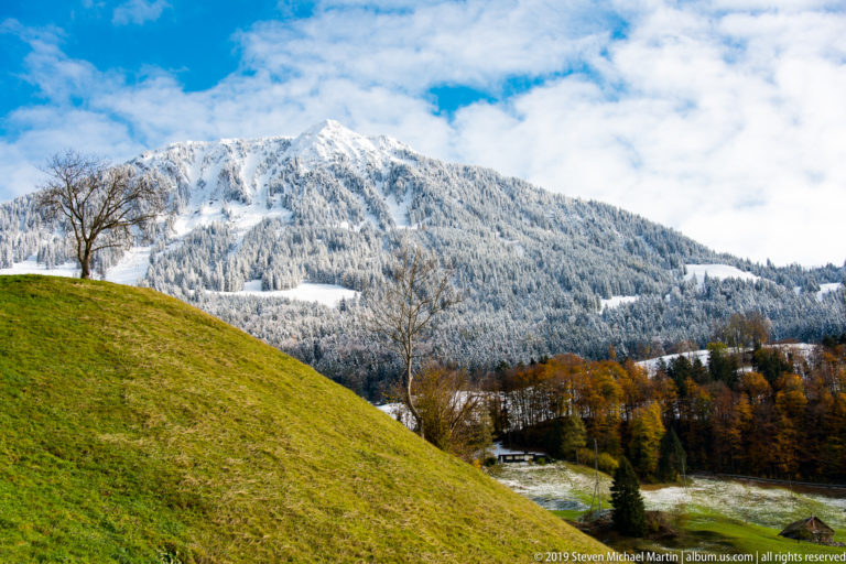 Green pasture below a snow-covered forested Alpine ridge under broken blue sky.