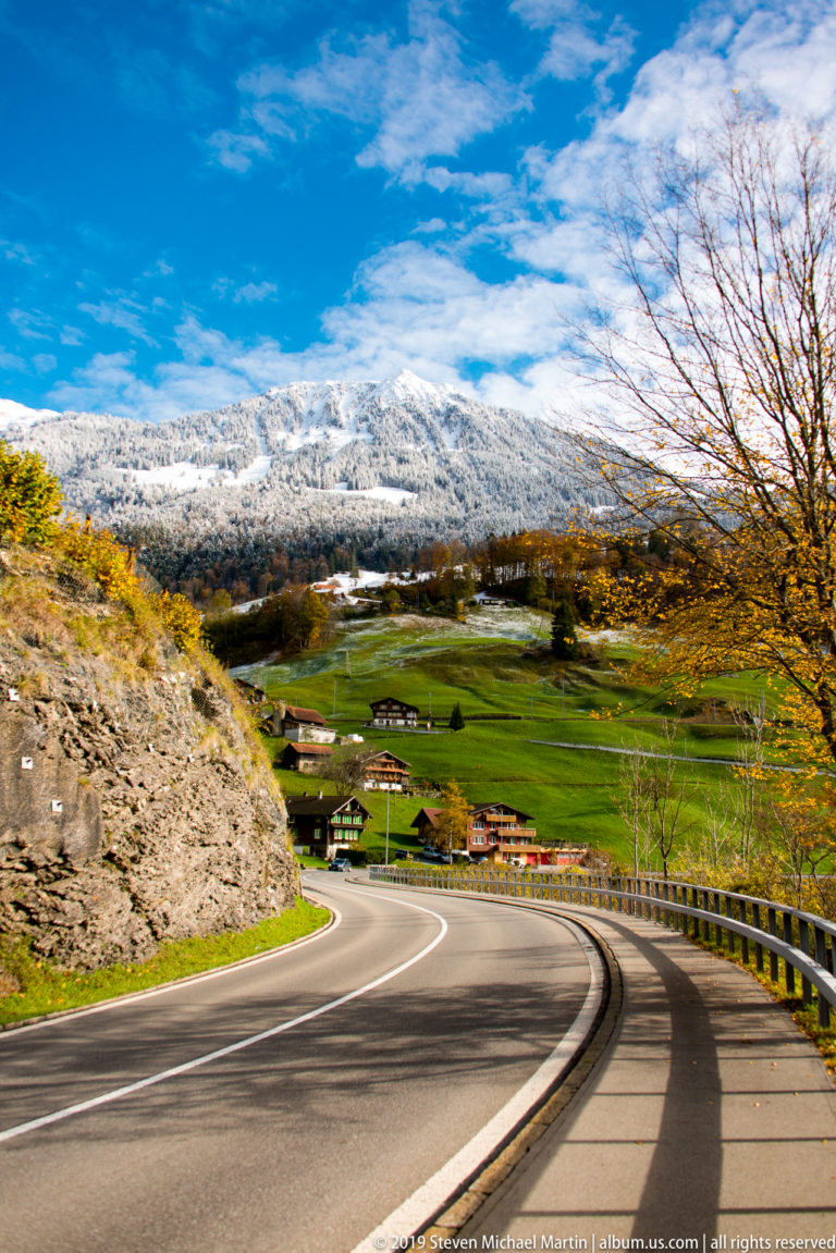 Curving two-lane road descending toward farmhouses and green fields beneath snowy mountains.