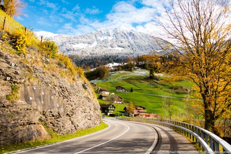 Road curves past a rock cut and golden tree toward Alpine houses and snowy slopes.