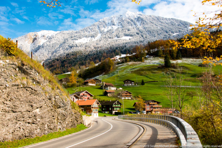 Winding road enters a chalet settlement below green fields and a snow-covered mountain.