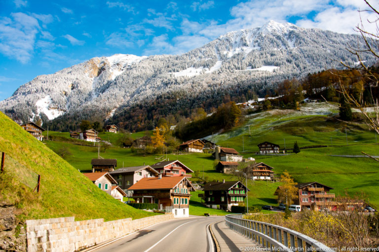 Roadside Alpine houses and scattered chalets beneath green slopes and a snowy ridge.