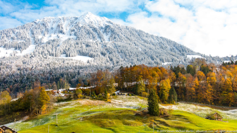 Green pasture and autumn trees rise toward a broad snow-covered Alpine mountain.