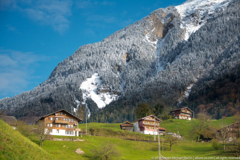 Chalet-style houses on green pasture beneath a steep, snow-streaked mountain.